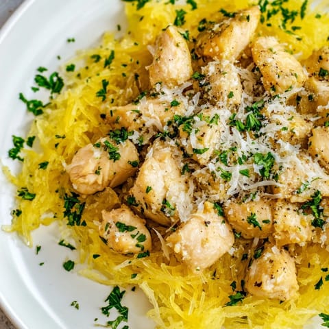 A close-up shows golden roasted spaghetti squash "noodles" topped with savory lemon garlic chicken, a sprinkle of Parmesan, and fresh herbs.  