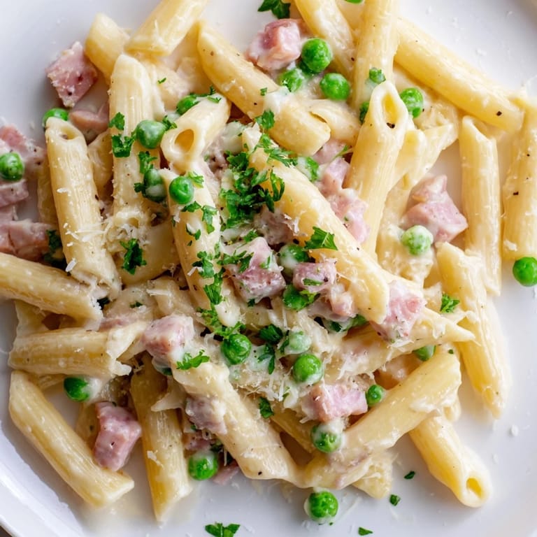 In a white bowl, Spring Pea & Ham Pasta showcases fork-tossed penne, ribbons of fresh parsley, and melted Parmesan ready for a hearty family dinner.